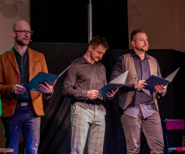 Na scenie trzech recytujących kabareciarzy trzymających w rękach niebieskie teczki 