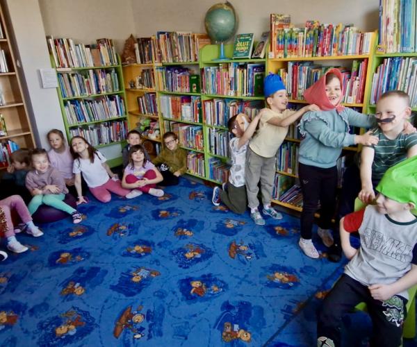 Dzieci trzymają się za plecy jeden za drugim w tle kilkoro dzieci siedzi na dywanie 